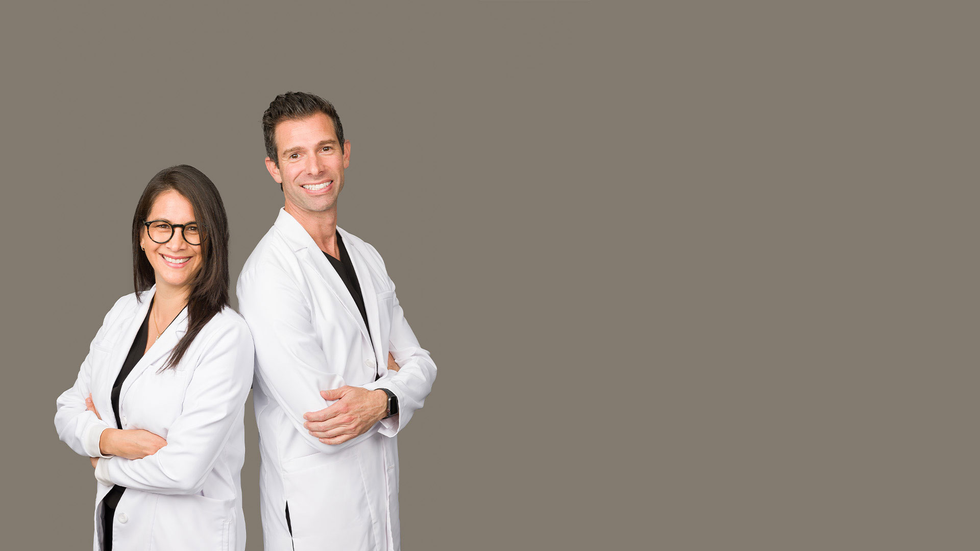 The image shows a man and a woman standing side by side, both wearing white lab coats, with the man on the left and the woman on the right. They are posing for the photo with one hand in their pocket and the other arm extended slightly away from their body.