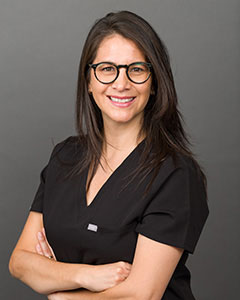 A woman in a black shirt, glasses, and a lanyard stands confidently with her arms crossed against a neutral background.