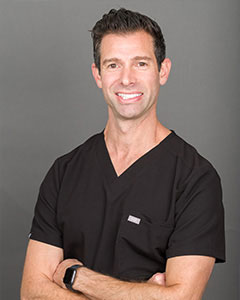 The image shows a man wearing a white lab coat, standing with his arms crossed in front of him. He has short dark hair and is smiling at the camera.