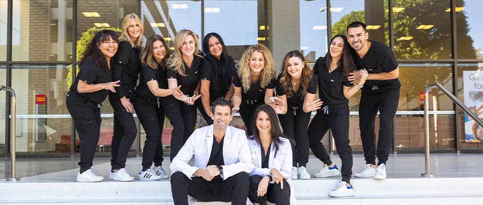 A group of smiling people in black scrubs posing for a photo, standing on a step outside a building with a glass door and a tree behind them.