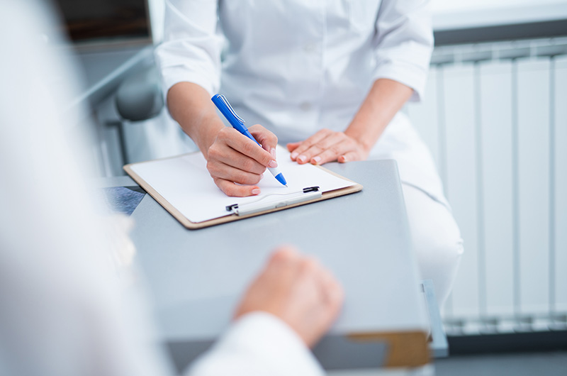 The image shows a person writing on a clipboard while seated at a table with another individual present, both appear to be in a professional setting.