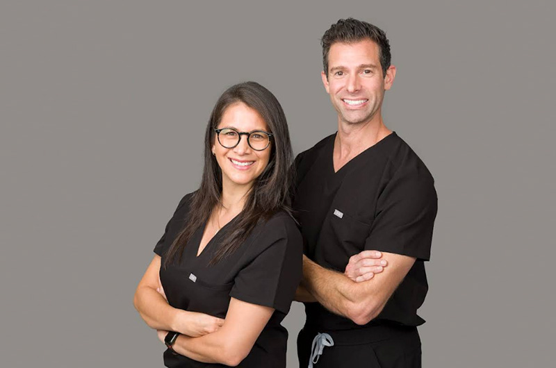 The image features two individuals posing together  the person on the left appears to be a woman wearing glasses, standing behind a man who is smiling and has his arms crossed. Both are dressed in professional attire, suggesting they may be healthcare workers, with the man wearing a stethoscope around his neck. They are set against a neutral background.