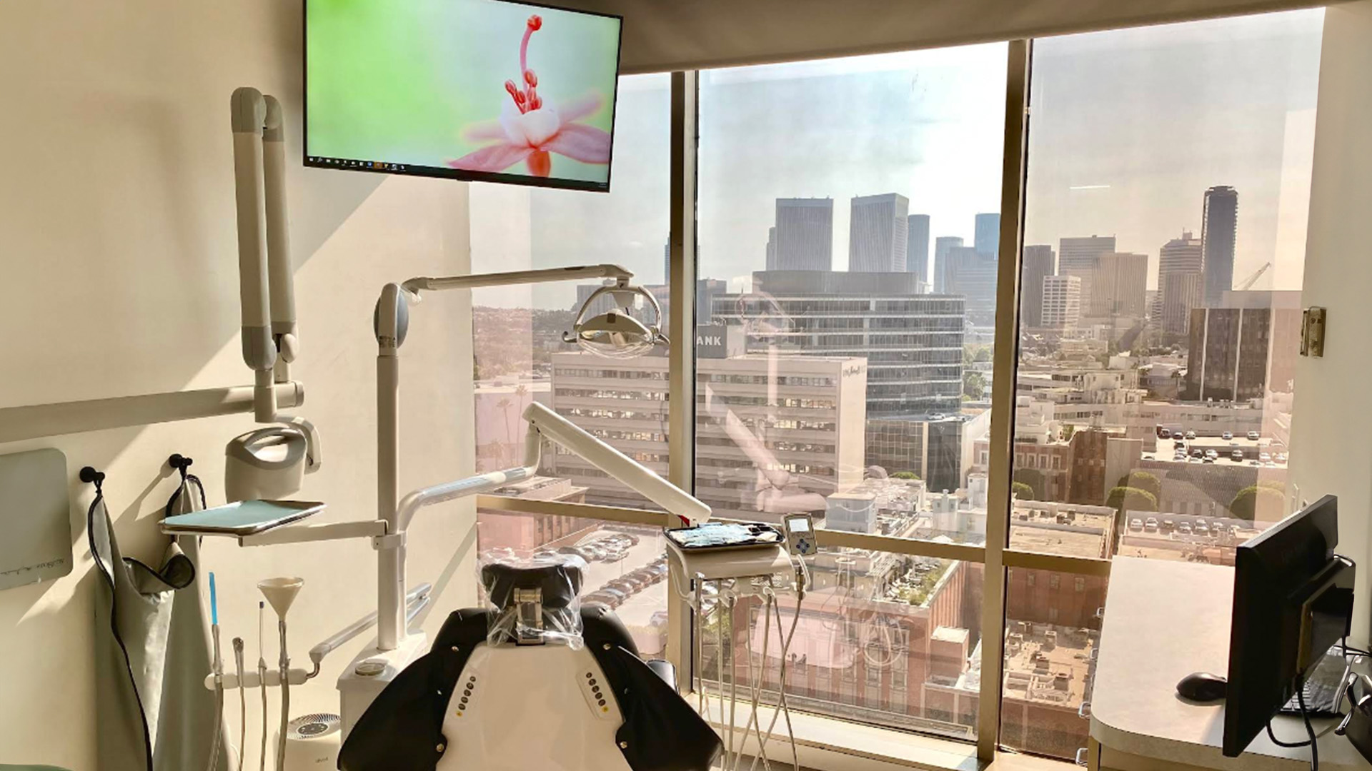 The image shows a room with dental equipment, including a chair, a television displaying an animated character, and a view of a city skyline through a large window.