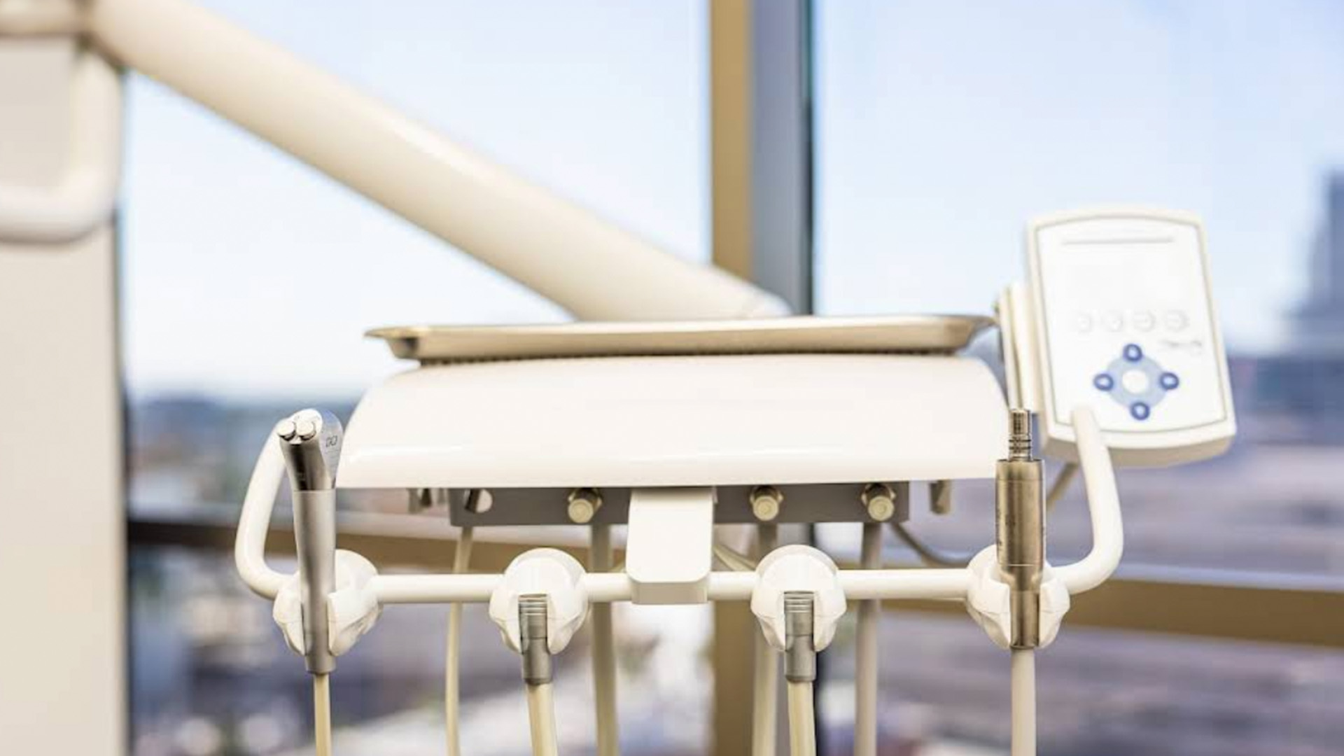 The image shows a modern dental chair with various equipment and screens attached to it, set against a window with an urban view outside.