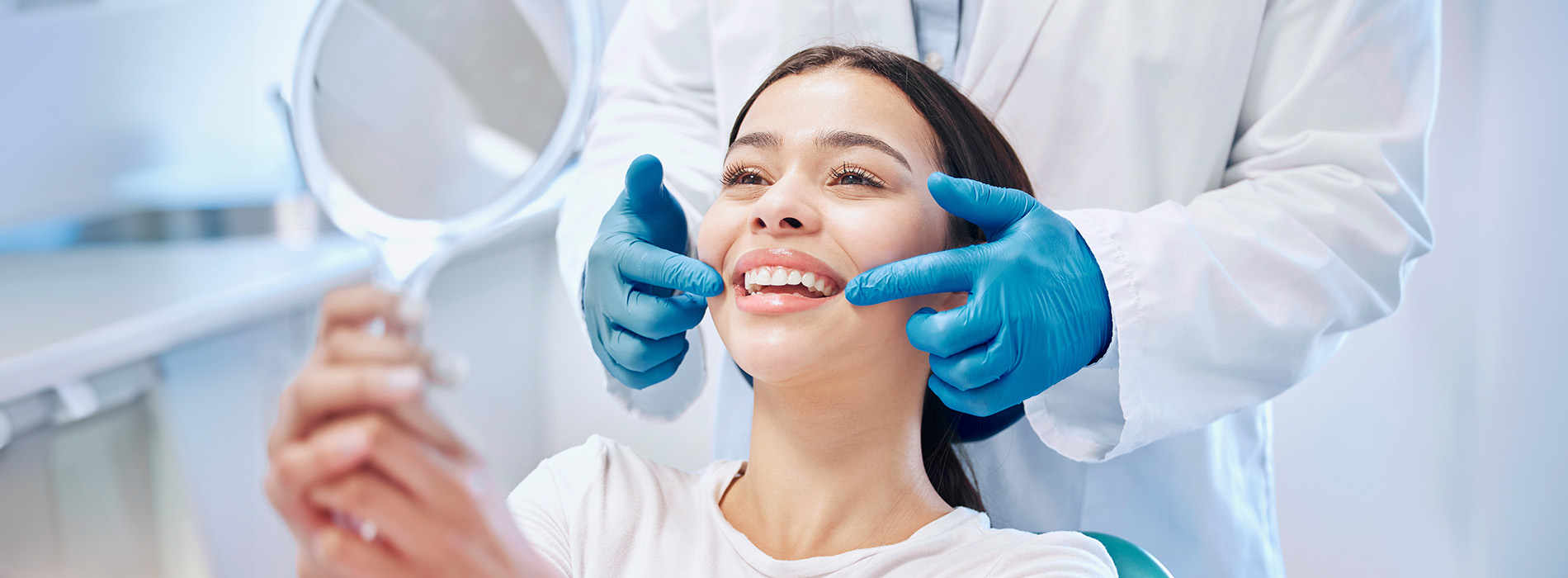 In the image, a woman is seated in a dental chair with her eyes closed, receiving dental treatment from a professional wearing blue gloves and a white lab coat, who is holding a mirror over her face.