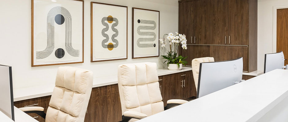 The image shows an office interior with a modern design, featuring a desk and chair setup, framed artwork on the wall, and a comfortable seating area.