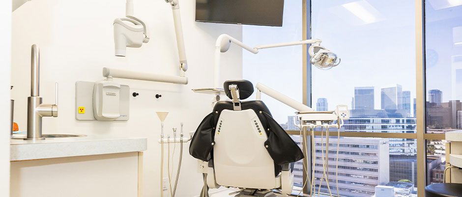 The image shows a modern dental office interior, featuring a dental chair with a black headrest and a digital display, a large window with city skyline view, a wall-mounted TV screen, dental equipment on the countertop, and a reception desk in the background.