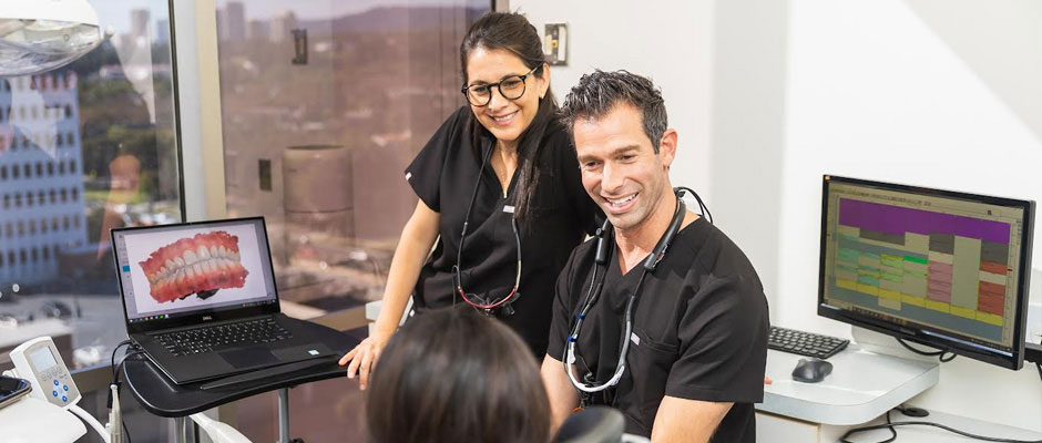 The image depicts a man and woman in a dental office, with the man seated at a computer desk and the woman standing beside him. They are both wearing white coats, suggesting they may be dental professionals.