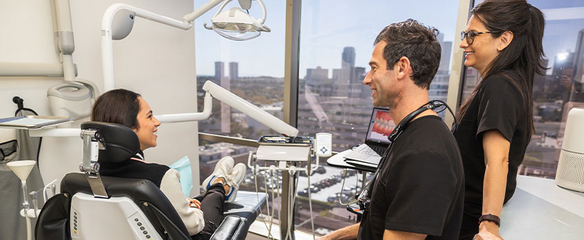 The image shows a dental office setting with a dentist and patient, featuring dental equipment and a view of the city through a large window.