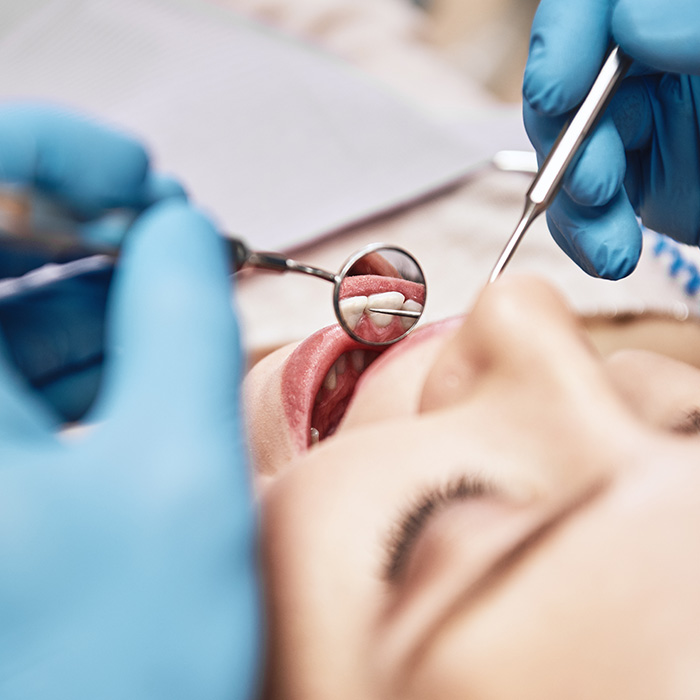 The image shows a person receiving dental treatment with a dental mirror being used to examine their teeth while they are seated in a dental chair, surrounded by medical equipment and a dental professional wearing gloves and a face mask.