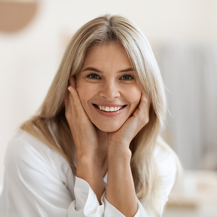 The image features a woman with blonde hair smiling at the camera, wearing a white top, with her hand on her cheek. She appears to be indoors, possibly in a home or office setting.