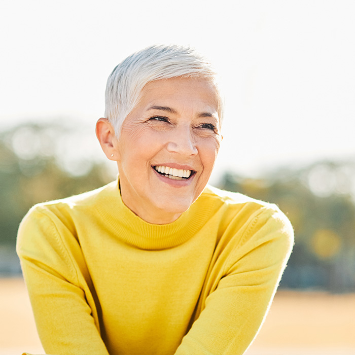 The image shows a woman with short hair smiling at the camera, wearing a yellow top and sitting outdoors during daylight.