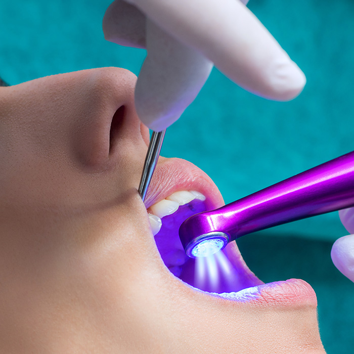 The image shows a person receiving dental care, with an electric toothbrush cleaning their teeth under the supervision of a dental professional wearing gloves and using a magnifying loupe.