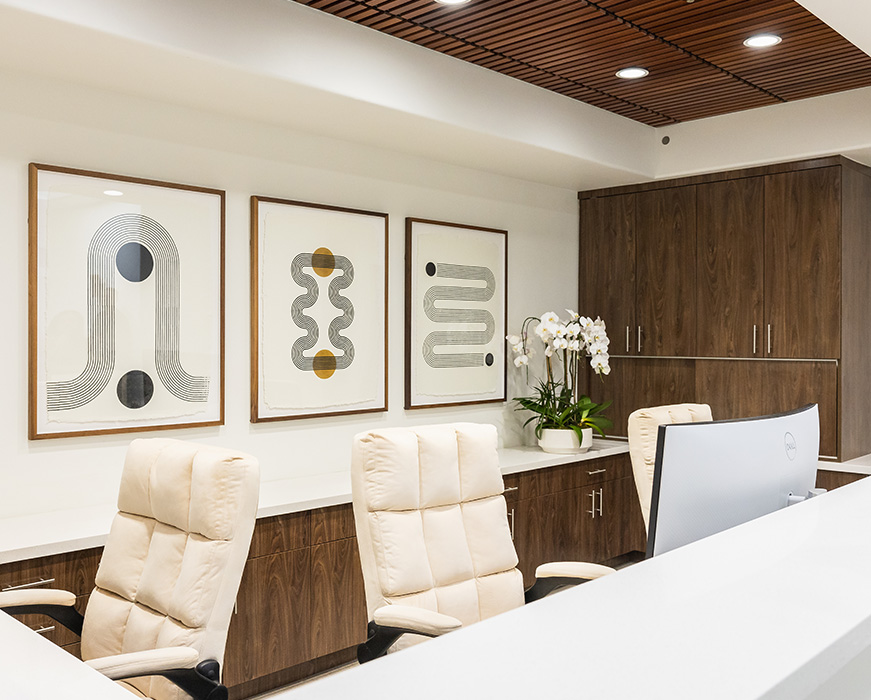 This is a photograph of an interior space featuring a modern office setting with a clean, minimalist design. The room has a large desk with chairs, a sleek countertop with a laptop, a contemporary kitchenette area with cabinets and appliances, and a wall displaying abstract artwork.