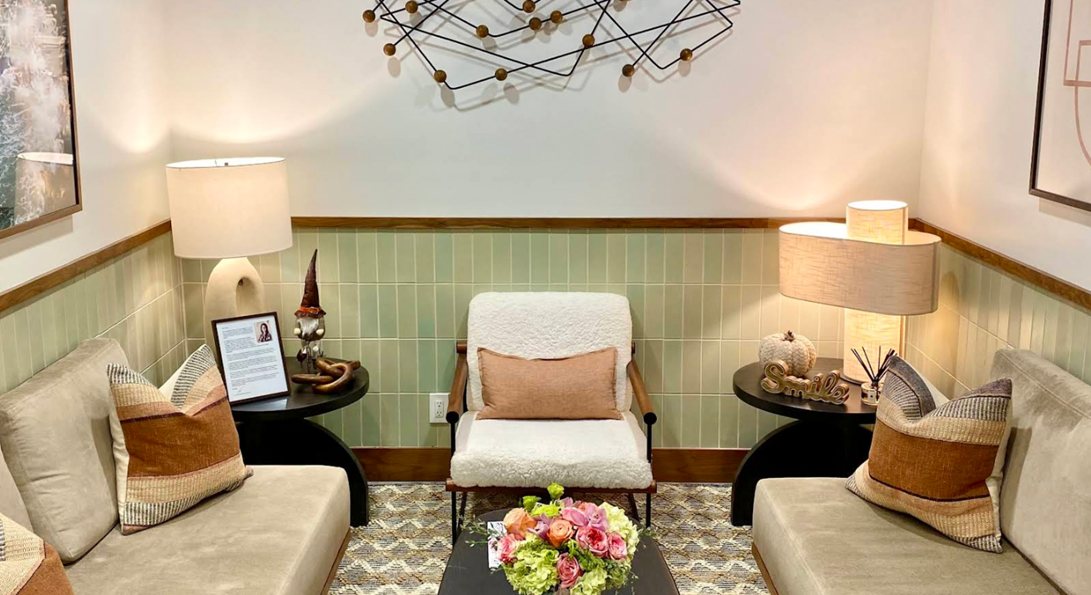The image shows an interior space with modern decor, featuring a seating area with two chairs and a small table, a wall-mounted light fixture, a framed picture on the wall, and a green accent tile above a chair rail.