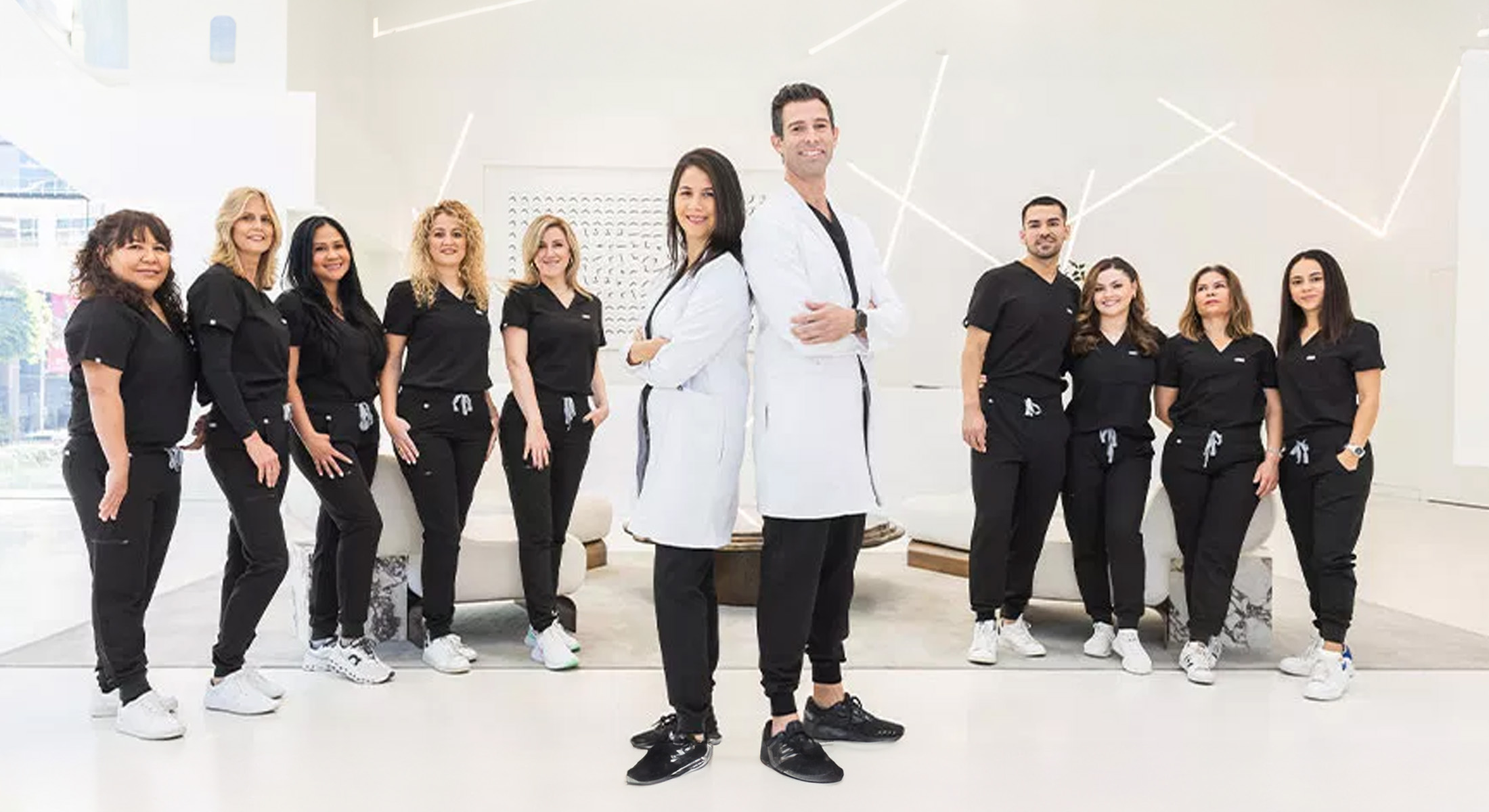 A group of people, including a man standing with his hands on hips, are posing for a photo together, likely at a formal event or professional setting, with some individuals wearing scrubs and others dressed in casual attire.