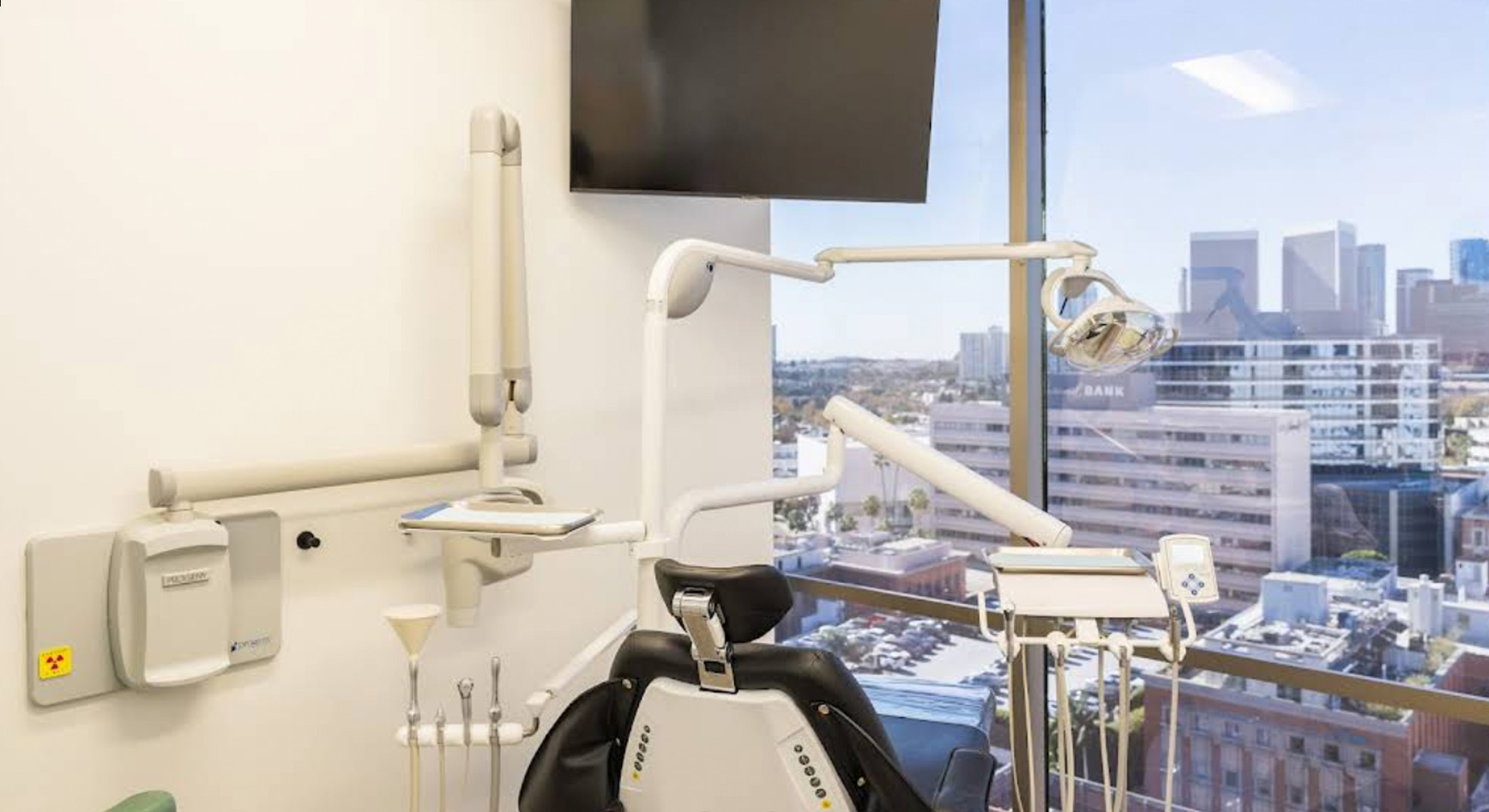 The image depicts a dental office with a cityscape visible through the window, featuring a city skyline under a clear blue sky during daylight hours.