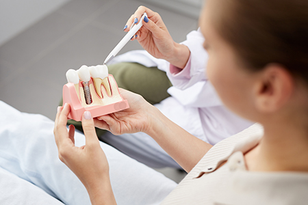 A dental hygienist holding a model tooth with a drill while standing in front of a patient in a dental chair.