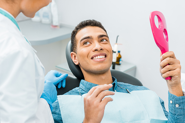 A man sitting in a dental chair with a smile on his face, holding a pink object that appears to be a dental device, while being attended to by a dental professional.