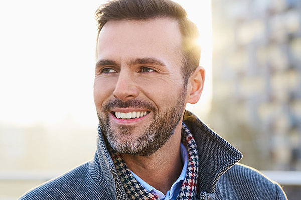 The image features a man with facial hair, smiling, and wearing a checkered scarf. He has short hair and is dressed in a casual style, standing outdoors during daylight.